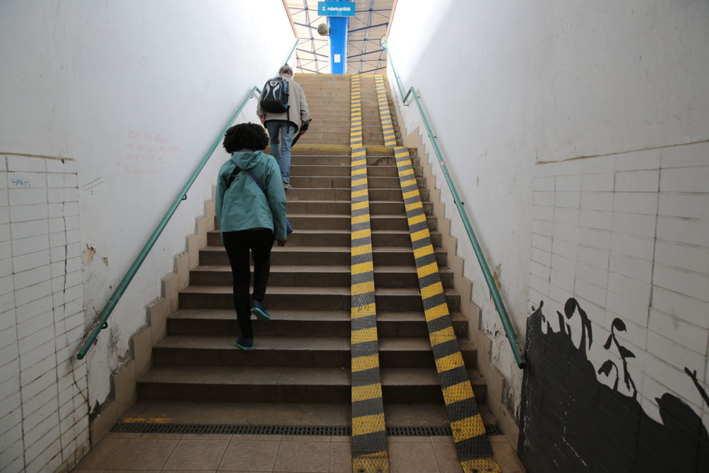 An image of a tall staircase covering at least two floors is visible in a public space. The backs of two people ascending the stairs are visible at left. No elevator or other automated technology is visible; instead, two thin tracks are laid over the right side of the staircase, about as far apart as the wheels on a wheelchair, making a very steep ramp.