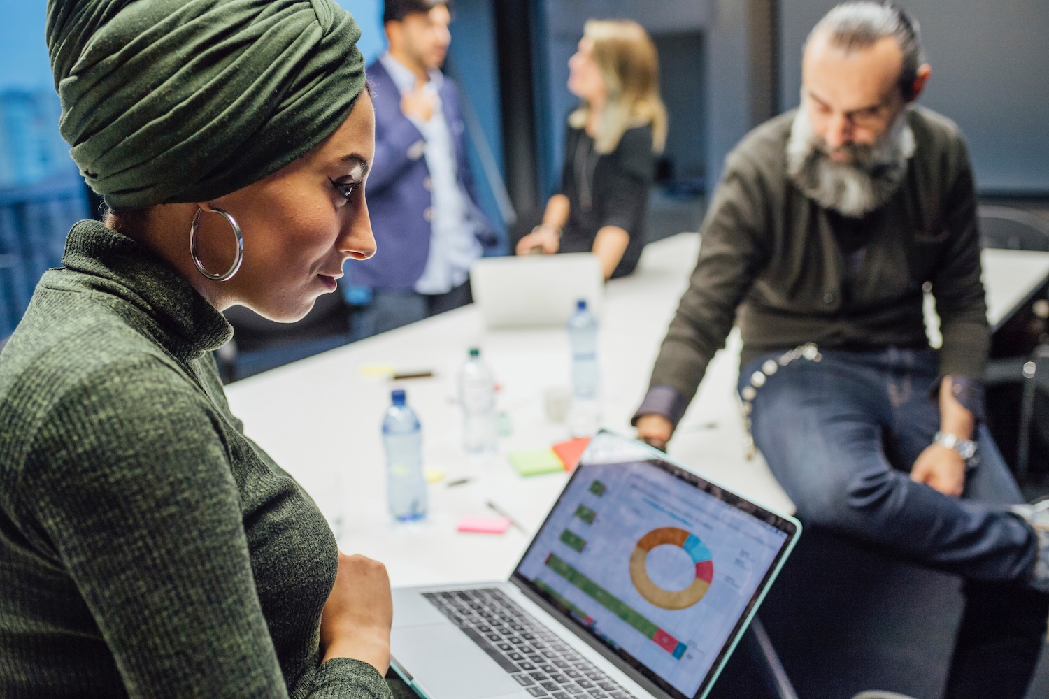 A woman in focus as people sit around a conference table, with a laptop open displaying a pie chart