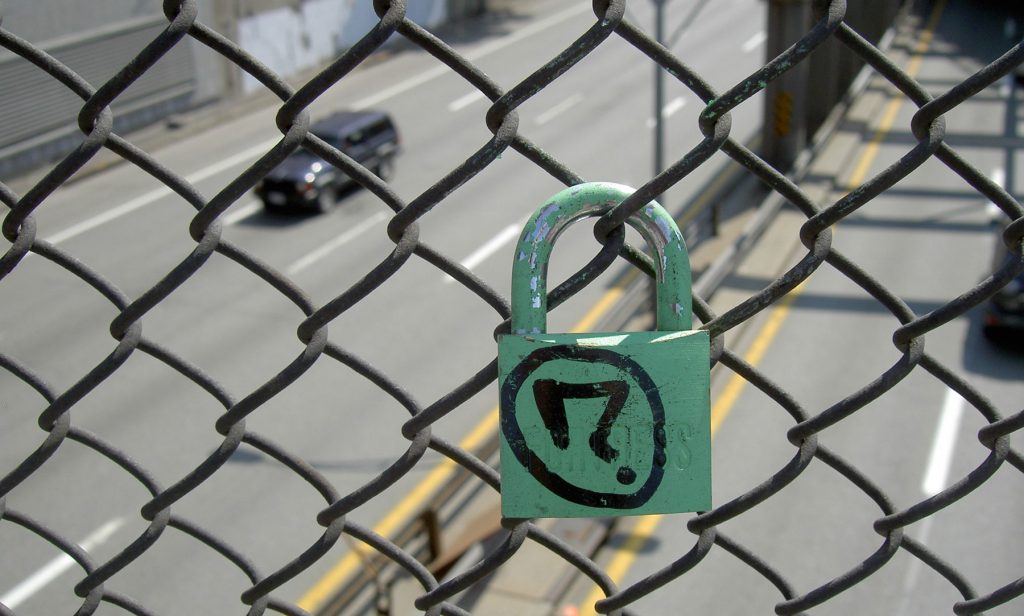 A green padlock is attached to a fence in front of a roadway.