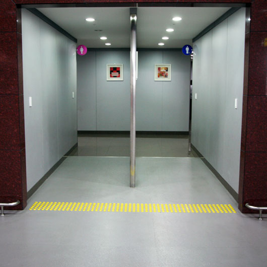 A restroom entrance with no doors. There are two corridors side-by-side, with a metal divider in the middle. In the upper corner near the back of both corridors, there's a small circular sign with a pictogram of a man or a woman, with a blue or pink background, respectively.