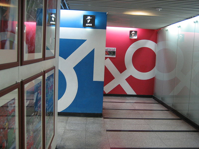 A bathroom entrance in Seoul, South Korea. The men's bathroom is denoted by the sex symbol for male in white on a blue wall, while farther down the hall is the female sex symbol in white on a reddish wall.