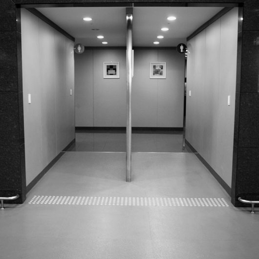 A restroom entrance with no doors. There are two corridors side-by-side, with a metal divider in the middle. In the upper corner near the back of both corridors, there's a small circular sign with a pictogram of a man or a woman. The image has been modified to be in black and white.