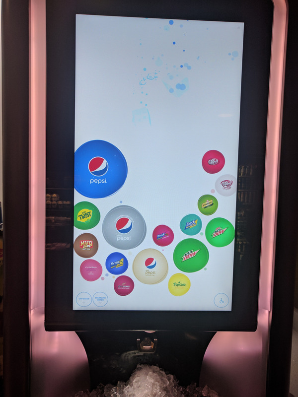 A closeup of a very tall touchscreen Pepsi dispenser. The various soda options are arrayed in a half-circle that stretches nearly two-thirds of the height of the screen, with some more popular brands much larger than others. An accessibility icon is in the lower-right corner of the screen.